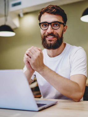Uzaktan kumandalı iş ve internet işlerinden memnun olan yetenekli erkek portresi kablosuz ağ ve dizüstü bilgisayarı para kazanmak için kullanıyor, gözlüklü zeki hippi adam kameraya bakıyor.