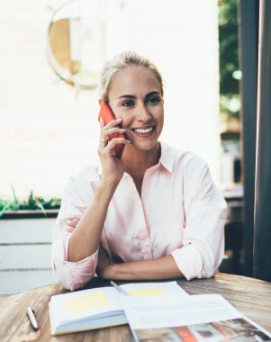 Nazikçe gülümseyen pembe tişörtlü genç sarışın masada oturuyor ve pencerenin yanındaki kafede akıllı telefondan sohbet ediyor.