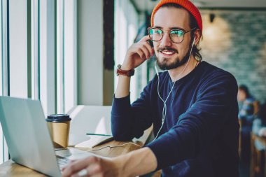 Optik gözlüklü olumlu erkek meloman, dost canlısı bir sesli mesajla kendini iyi hissediyor, kafeteryada dizüstü bilgisayarla oturan ve kulaklıkla müzik listesi dinleyen mutlu hipster adam.