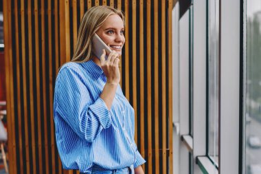 Elinde not defteri olan çekici hipster öğrenci akıllı telefon cihazıyla konuşuyor ve pencereden dışarı bakıyor. Modern çalışma alanında duruyor. Telefon konuşması sırasında gülen genç bir kadın.