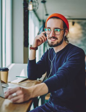 Optik gözlüklü olumlu erkek meloman, dost canlısı bir sesli mesajla kendini iyi hissediyor, kafeteryada dizüstü bilgisayarla oturan ve kulaklıkla müzik listesi dinleyen mutlu hipster adam.