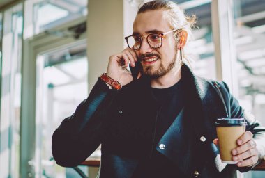 Mutlu erkek milenyum zamanı kahve dükkanında vakit geçirip iş yerinde buluşma zamanını konuşmak için arkadaşını arıyor. Hücresel iletişim yoluyla elinde kafein içeceği olan pozitif hipster adam.