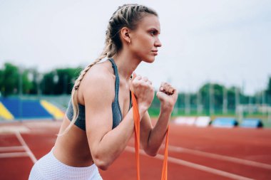 Açık hava stadyumunda kol kasları açmak için kauçuk jimnastik bandı geren uzun sarı saçlı, konsantre spor kadın.