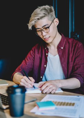 Gözlüklü zeki Asyalı genç adam dizüstü bilgisayarda webinar izlerken metin bilgileri yazıyor. Japon hippi öğrenci modern çalışma alanında dijital bilgisayarın başında ödev yapıyor.