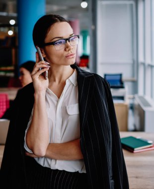 Gözlüklü ciddi bir kadın ofisinden uzaklaşıyor iş için cep telefonuyla konuşuyor resmi kıyafet giymiş kendine güvenen bir iş kadını operatörle akıllı telefon konuşmalarıyla bankacılık dengesini kontrol ediyor.