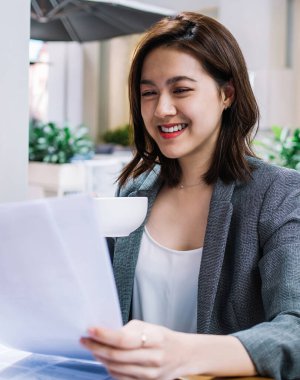 İş merkezindeki kafede dizüstü bilgisayarla masada oturan ve fincandan kitap okuyup içerken elinde belgelerle gezen mutlu kadın cep telefonu çalışanı.
