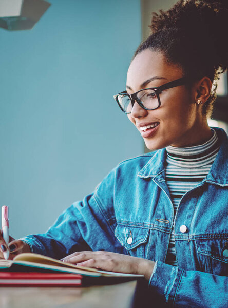 Cheerful female blogger feeling excited from creating interesting idea for content to next publication, happy dark skinned student spending time in cafeteria for preparing information for school test