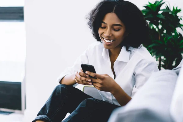 Cheerful dark skinned female blogger laughing during online chat with followers on phone using home internet.Positive african american young woman reading good news on smartphone relaxing on couch