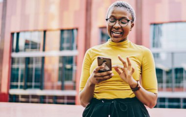 Mutlu Afro-Amerikalı kadın cep telefonuyla internet mağazasında satıştan heyecan duyuyor, neşeli esmer tenli hippi kız şehrin arka planında durmuş içeriğiyle gülüyor.