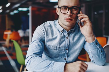 Ofisteki modern akıllı telefon cihazını ararken kameraya bakan şık gözlüklü ciddi bir genç adamın portresi. Kot gömlek giyen dalgın hippi öğrenci cep telefonuyla konuşuyor.