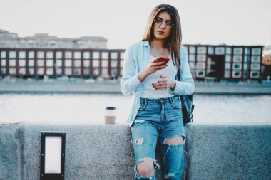 Kot pantolon giymiş genç bir kadının portresi, internet üzerinden mesajlaşırken kameraya bakarak kamuya açık 4G internet bağlantısını kullanıyor. Şehir merkezinde duran çekici hippi kız turist.