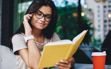 Vizyon düzeltmesi için gözlüklü olumlu hippi kız kafede boş zamanlarında en çok satan yeni kitabı okudu, edebiyata düşkün beyaz bir kadın kahve molası üzerine ilginç bir kitapla tatmin oldu.