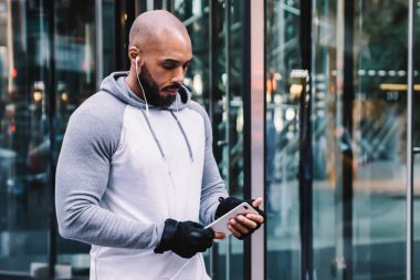 Spor giyim sektöründe kel Afrikalı Amerikalı adam akıllı telefon kullanıyor ve modern ofis binasının kapısında durup müzik dinliyor.