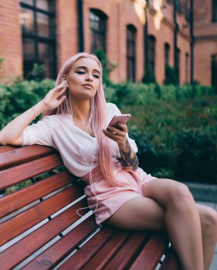 Beyaz gömlekli, pembe şortlu, uzun pembe saçlı, akıllı telefondan müzik dinleyen, tuğla binanın karşısındaki bankta oturan genç bir bayan.