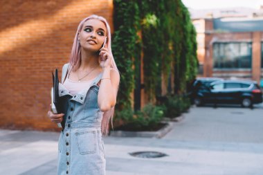 Gülümseyen ve arkasını kollayan defterleri olan neşeli genç bir kadın kaldırımda dikilip telefonlarına cevap veriyor.