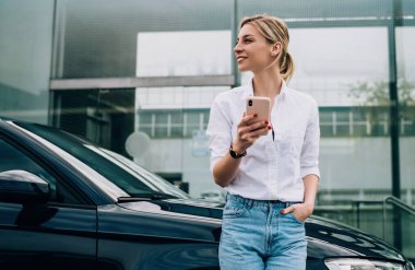 Siyah arabaya yaslanıp akıllı telefondan mesajlaşırken günlük kıyafetlerle gülümseyen neşeli sarışın kadın sürücü.