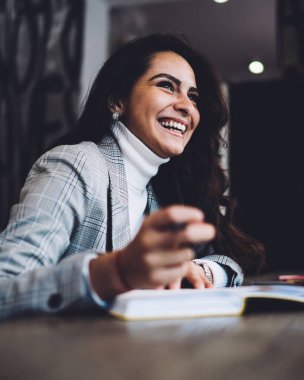 Kahkahalarla gülen, uzun siyah saçlı, günlük kıyafetlerle neşeli, kafede dizüstü bilgisayarla oturup notlar alan bir kadın.