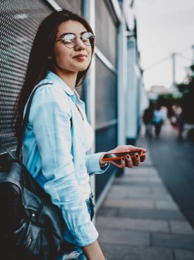 Gündelik giyinmiş çekici hippi kız portresi şehir ortamında akıllı telefonla dururken kameraya bakıyor. Sırt çantalı, şık bir kadın blogcu.
