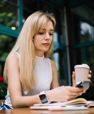 Kolsuz balıkçı yaka ve dijital saat takan genç sarışın kadın, gün ortasında telefon okumaya ve kahve dükkanının önünde kahve içmeye odaklanmış.