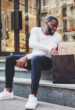 Uzun kollu tişörtlü, gülümseyen hippi adam alışveriş çantalarına bakıyor, alışverişleri kontrol ediyor, Afro-Amerikan erkek iyi bir indirimle giyim mağazasından kıyafet almaktan mutlu.