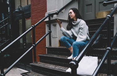 Kot pantolon ve rahat süveter giyen güzel yetişkin bir kadın New York 'un caddesinde gezerken alışveriş çantalarını merdivenlere koyuyor.