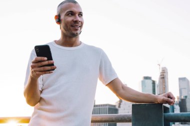 Beyaz gömlekli, kulaklıklı, akıllı telefonlu, metalik çit ve nehrin yanında duran kısa saçlı genç adam şehrin arka planına yaslanmış.