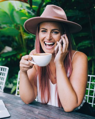 Güler yüzlü, şapkalı, akıllı telefondan konuşan ve kahve içen bir kadın tropik yaz gününde dizüstü bilgisayarla birlikte açık terasta oturuyor.