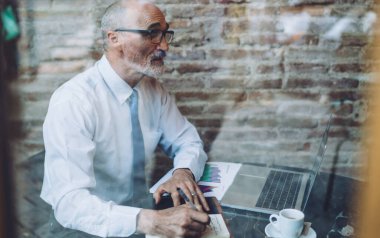 Gözlüklü, son sınıf kel, resmi kıyafetler giymiş, kafede dizüstü bilgisayarını incelerken not alan adama bakın.