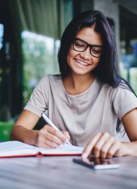 Kafkasyalı olumlu kadın cep telefonundan bildirimi kontrol ederken not defterine bilgi yazıyor, neşeli genç bayan blogcu takipçilerle akıllı telefon aracılığıyla sohbet ediyor ve kafede makaleler yaratıyor.