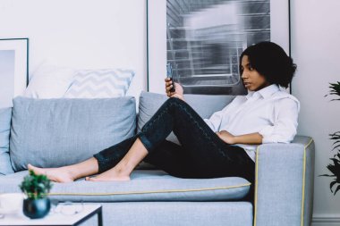 Afro-Amerikan tarzı genç bir kadın rahat bir kanepede uzanıyor ve akıllı telefon cihazıyla sosyal ağlarda bildirim okuyor. Ev internet bağlantısını kullanıyor. Kadın blogcu telefonda sohbet ediyor.