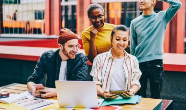 Gündelik giyinmiş çeşitli gençler modern akıllı telefondan komik şeyler izleyip dışarıda işbirliği yaparken dizüstü bilgisayarlarıyla oturuyorlar. 4G internet kullanan çok kültürlü hipster çocuklar.