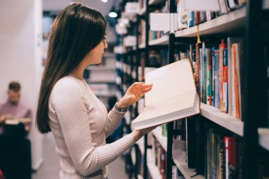 Kitap rafının önünde duran ve elinde kitap tutan güzel, ince bir kadının yan görüntüsü.