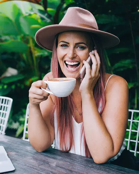 Güler yüzlü, şapkalı, akıllı telefondan konuşan ve kahve içen bir kadın tropik yaz gününde dizüstü bilgisayarla birlikte açık terasta oturuyor.