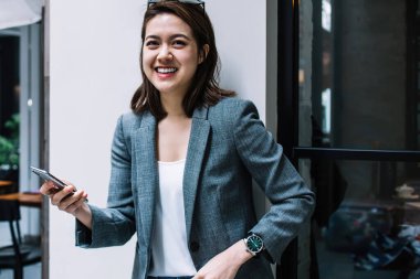 Genç, çekici, güzel, neşeli, resmi giyinmiş, akıllı telefon kullanan, duvara yaslanmış, caddede kameraya bakan bir kadın.