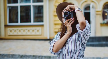Arka planda fotoğraf çeken yetenekli kadın fotoğrafçı şehir ortamlarında dikiliyor, modaya uygun şapka ve giysili beyaz kadın şehirde fotoğraf çektirip eğlendiriyor.