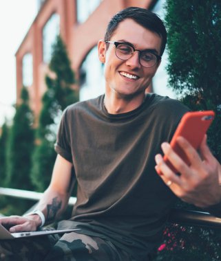 Görme düzeltmesi için gözlüklü neşeli erkek öğrenci akıllı telefon cihazıyla gülerek kampüste modern laptopla otururken, dışarıda serbest çalışan pozitif bir serbest oyuncu tarafından komik bir mesaj aldı.