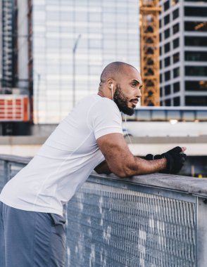 Spor giyim giyim giymiş kel sakallı bir zenci şehir merkezindeki ahşap iskelede korkuluklara yaslanıp kulaklıkla müzik dinliyor.