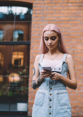 Dövmeli, uzun pembe saçlı, kot elbiseli modern odaklı kadın. Şehir caddesinde cep telefonundan mesajlaşıyor.