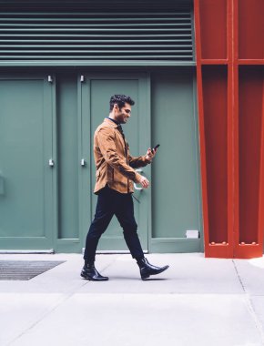 New York caddesindeki binaya karşı şık kıyafetler içinde yürüyen ve akıllı telefondan yazan gülümseyen genç bir erkek.
