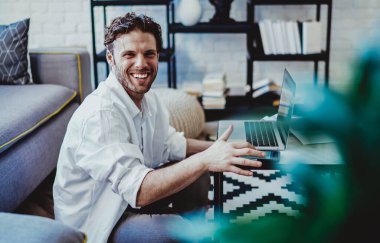 Neşeli beyaz erkek serbest çalışan, modern apartman dairesinde mesafesinin tadını çıkaran mutlu hippi adam, online kazançlarından memnun, dizüstü bilgisayar ve kablosuz internet üzerinden bloğundaki içeriği paylaşıyor.