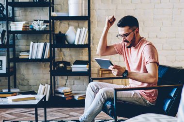 Modern dairede rahat bir sandalyede oturan genç hippi modern dijital tabletle online oyun oynarken zaferi kutluyor ve inanılmaz haberler okuyor.