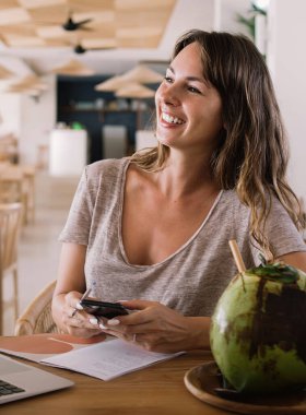 Çevrimiçi iletişim sırasında mutlu, neşeli bir kadın kendi blogundan seyahat serbest yaşam tarzıyla ilgili web takipçileriyle gülüyor. Egzotik hindistan cevizi içeceğiyle masada oturan yetenekli bir kadın.