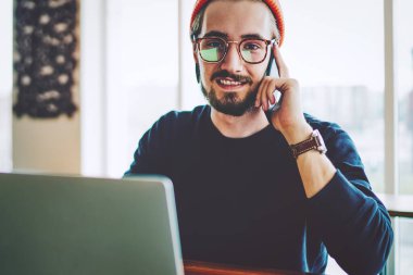 Akıllı telefon görüşmeleri sırasında kameraya gülümseyen gözlüklü başarılı erkek öğrencinin portresi, modern netbook ile aynı masada oturan pozitif genç adam.