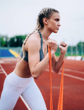 Açık hava stadyumunda kol kasları açmak için kauçuk jimnastik bandı geren uzun sarı saçlı, konsantre spor kadın.