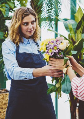Profesyonel kadın bahçıvan kendi botanik serasından taze çiçek satıyor, önlüklü satıcı müşteriye iş sırasında cam evde bitkiler yetiştirirken ipek buket sipariş ediyor.