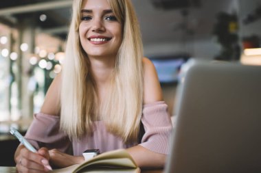 Açık pembe elbise giyen, gülümseyen ve kameraya bakan genç bir sarışın. Not defterine yazarken ve laptopla kafede otururken.