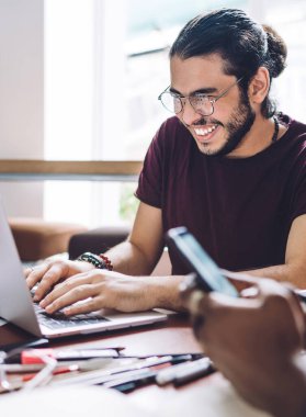 Gözlüklü neşeli bir adam dizüstü bilgisayarını kullanıyor. Not defteriyle oturuyor ve grup arkadaşlarıyla gülüyor.