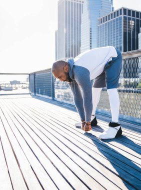 Spor giyim kuşamında atletik kel Afrikalı Amerikalı arka planda modern şehir ve nehirle koşmadan önce bacaklarını esnetmeye çalışıyor.