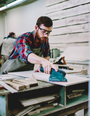 Önlük ve güvenlik gözlükleriyle sakallı katılımcı profesyonel marangoz atölyesinde masa üzerindeki ahşap kalası cilalamak için de kullanma kılavuzu kullanıyor.