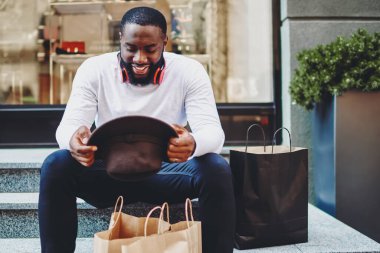 Alışveriş saatinden sonra merdivenlerde dinlenen, şık şapkalara bakan ve bir şeyler satın aldığı için kendini iyi hisseden, kağıt torbalı mutlu hippi erkek.